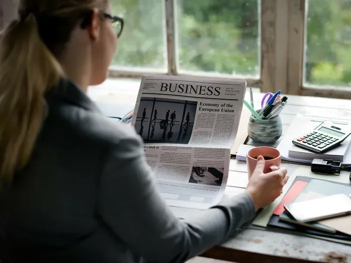 Professional woman reading business newspaper at office desk with coffee mug, laptop, and stationery items